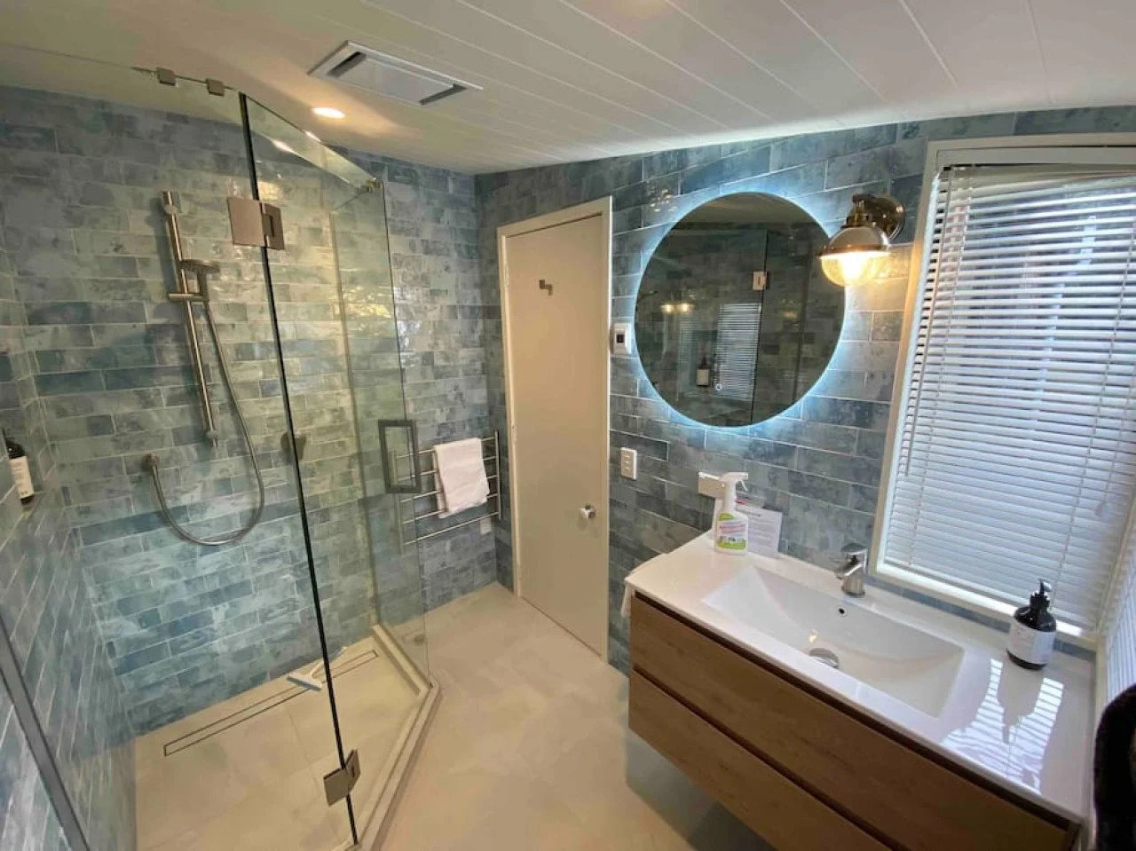 Modern bathroom with gray brick walls, a glass-enclosed shower area, a round backlit mirror, a white sink with wooden cabinet, a window with blinds, and a wall sconce light.