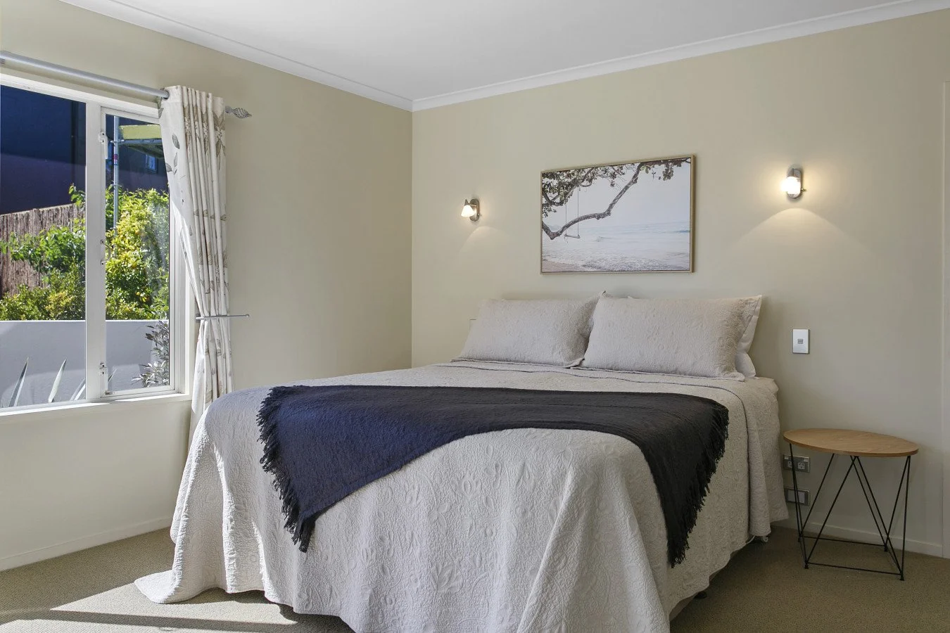 Bedroom with a bed covered in a cream quilt and a dark throw blanket, a nightstand, a painting of a tree and beach, and a window with curtains showing greenery outside.