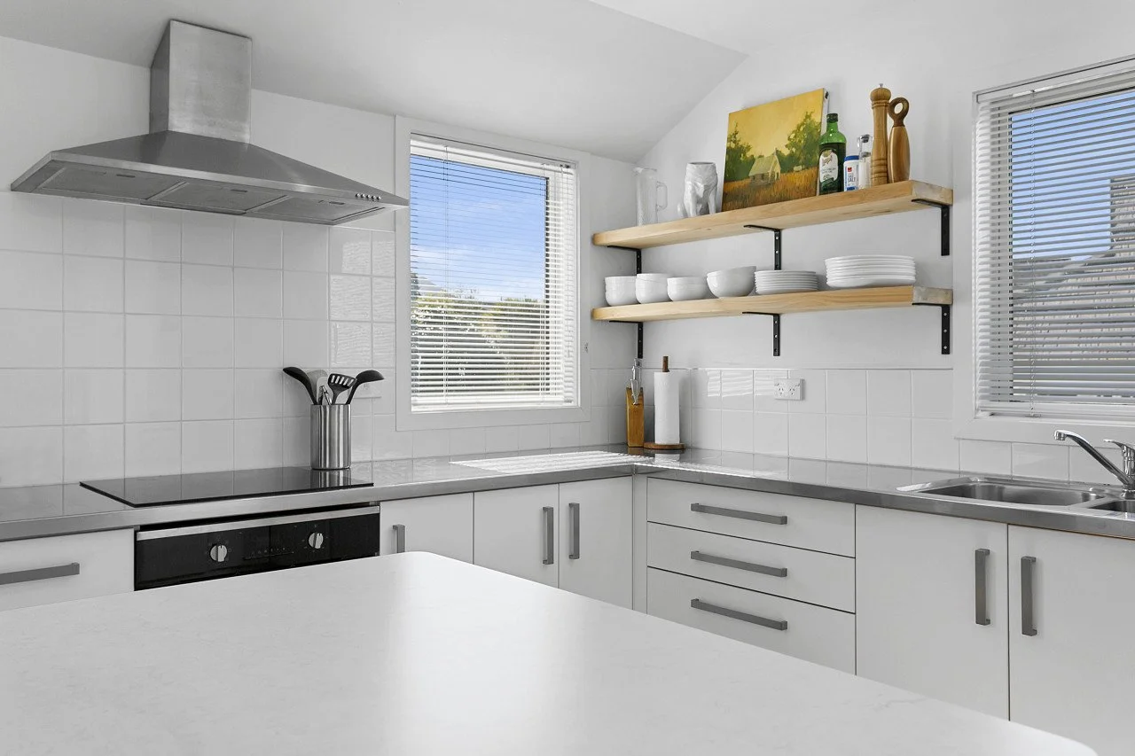 Modern white kitchen with open shelving, stainless steel appliances, and two windows with blinds