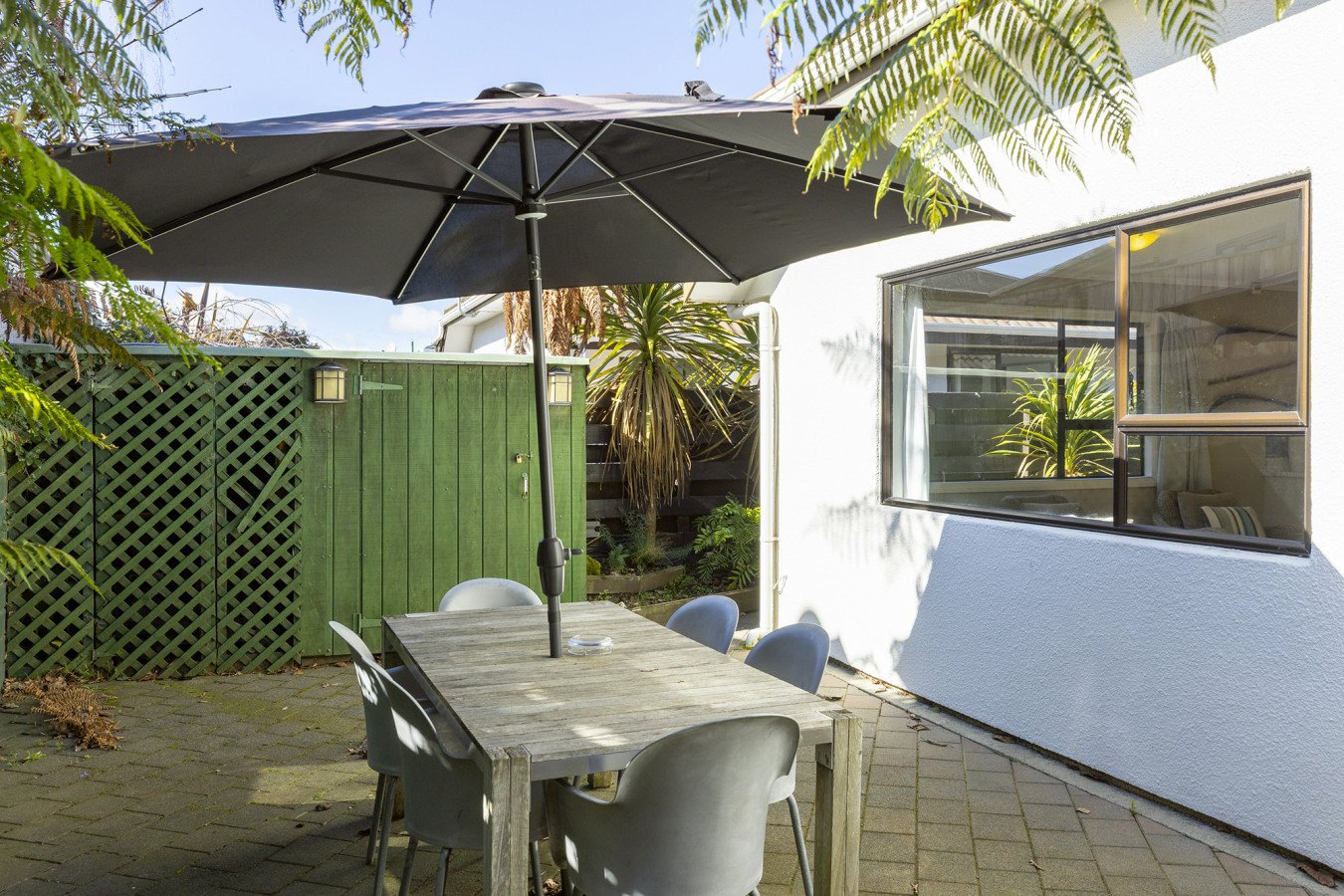 Outdoor patio with a wooden table, four gray chairs, black umbrella, green fence, garden plants, and a white house wall with a large window.