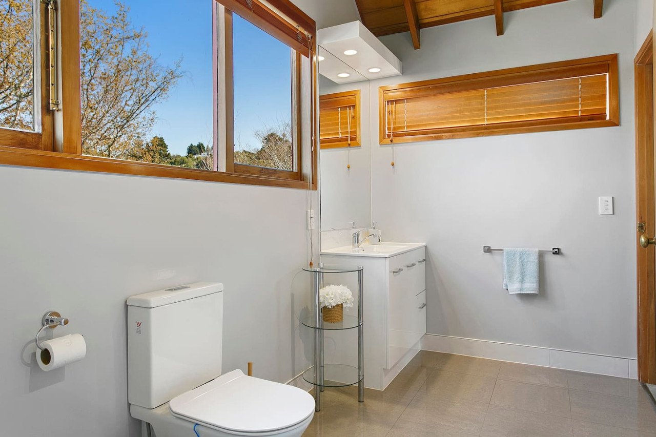 Bathroom with white walls, wooden window blinds, a toilet, a small white sink vanity, a towel bar with a blue towel, and a small round glass shelf with flowers.