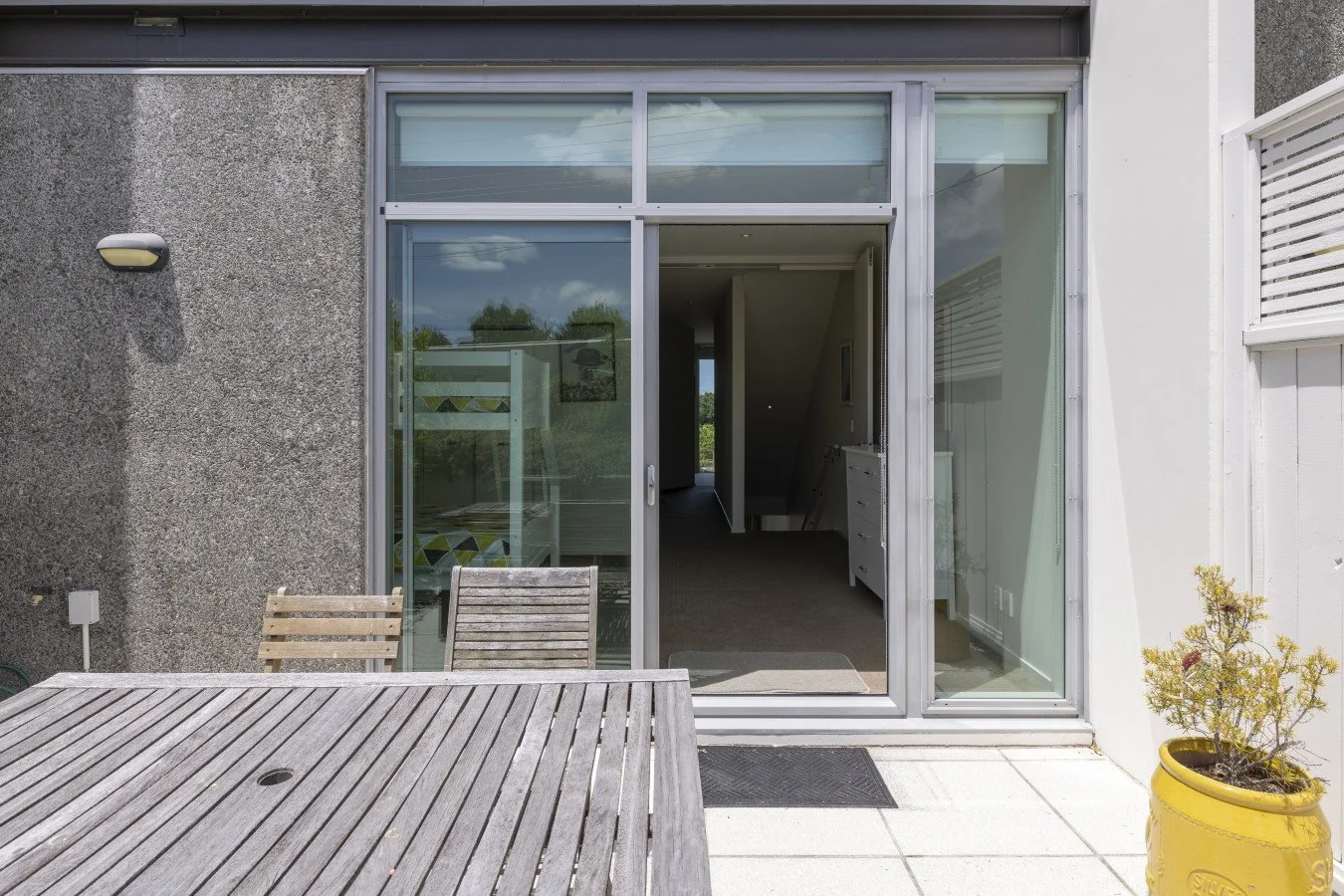 View of a modern house patio with a wooden table, two wooden chairs, a potted plant in a yellow pot, and a sliding glass door leading into the house.