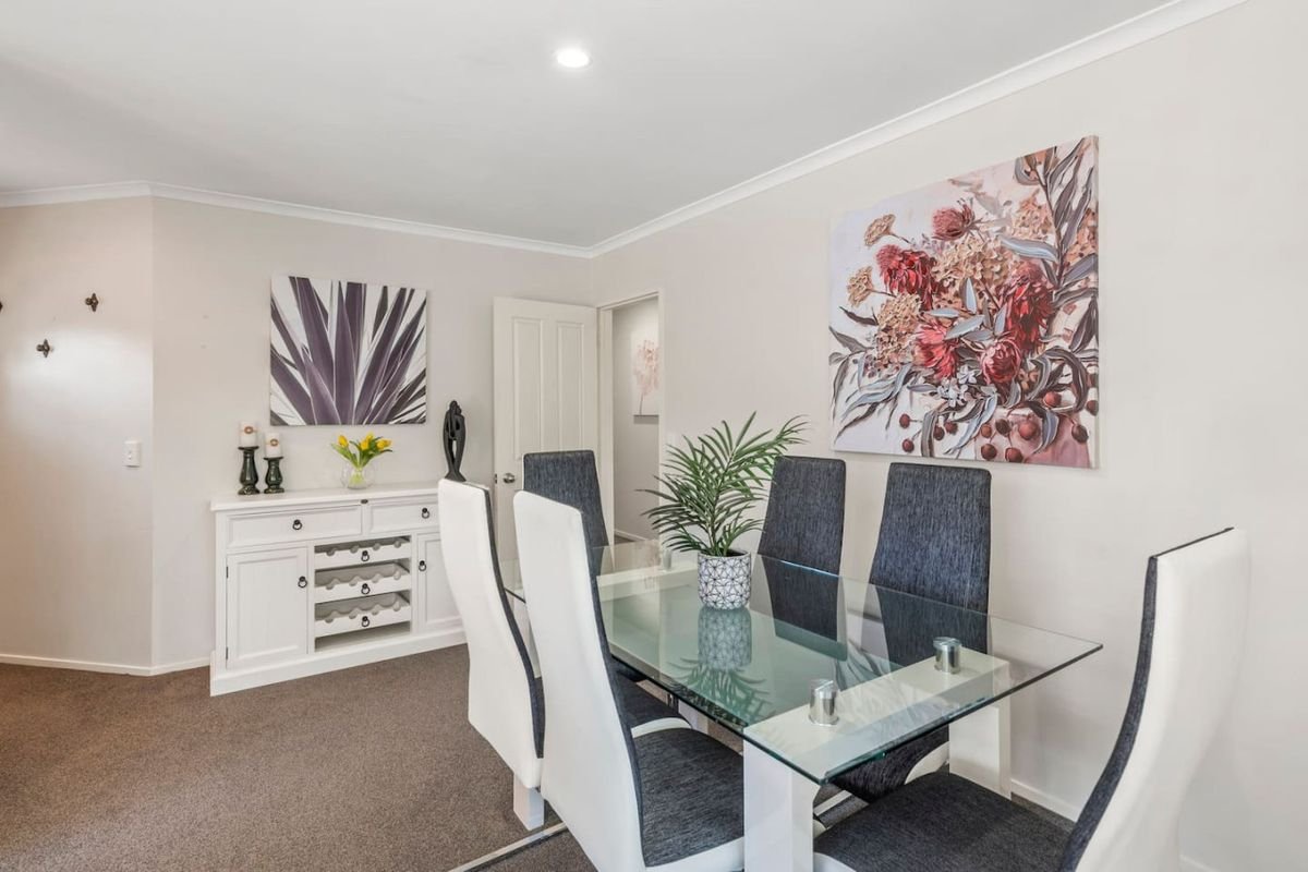 Dining room with a glass-top table surrounded by six high-back chairs, a white sideboard with decorative items, artwork on the walls, and a potted plant in the center of the table.
