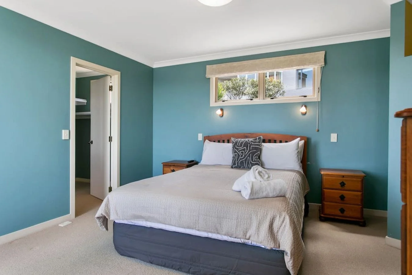 A bedroom with blue walls, a bed with beige bedding, pillows, and towels, alongside wooden nightstands and a window above the bed.