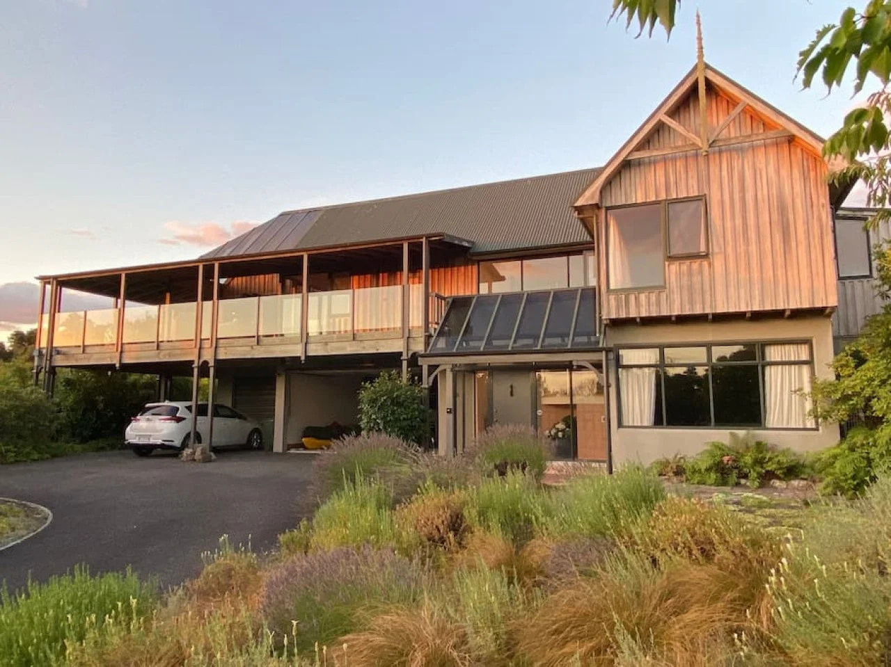 A two-story house with a mix of wooden and concrete exterior, featuring a large glass window, a balcony, and a driveway with two cars parked underneath. There are plants and bushes in front of the house.