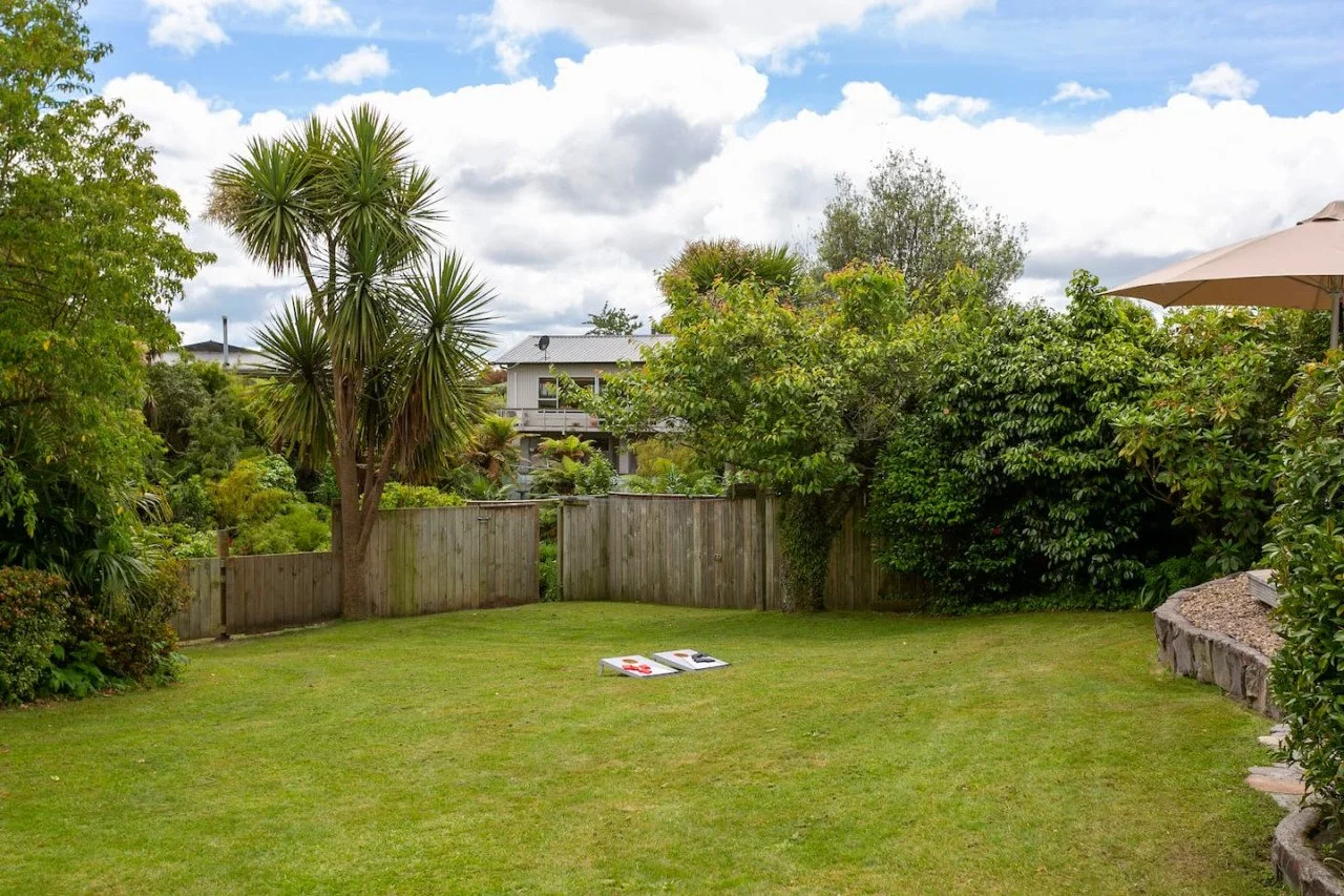 A backyard with green grass, surrounded by trees and bushes, with a wooden fence, and a house in the background under a cloudy sky.