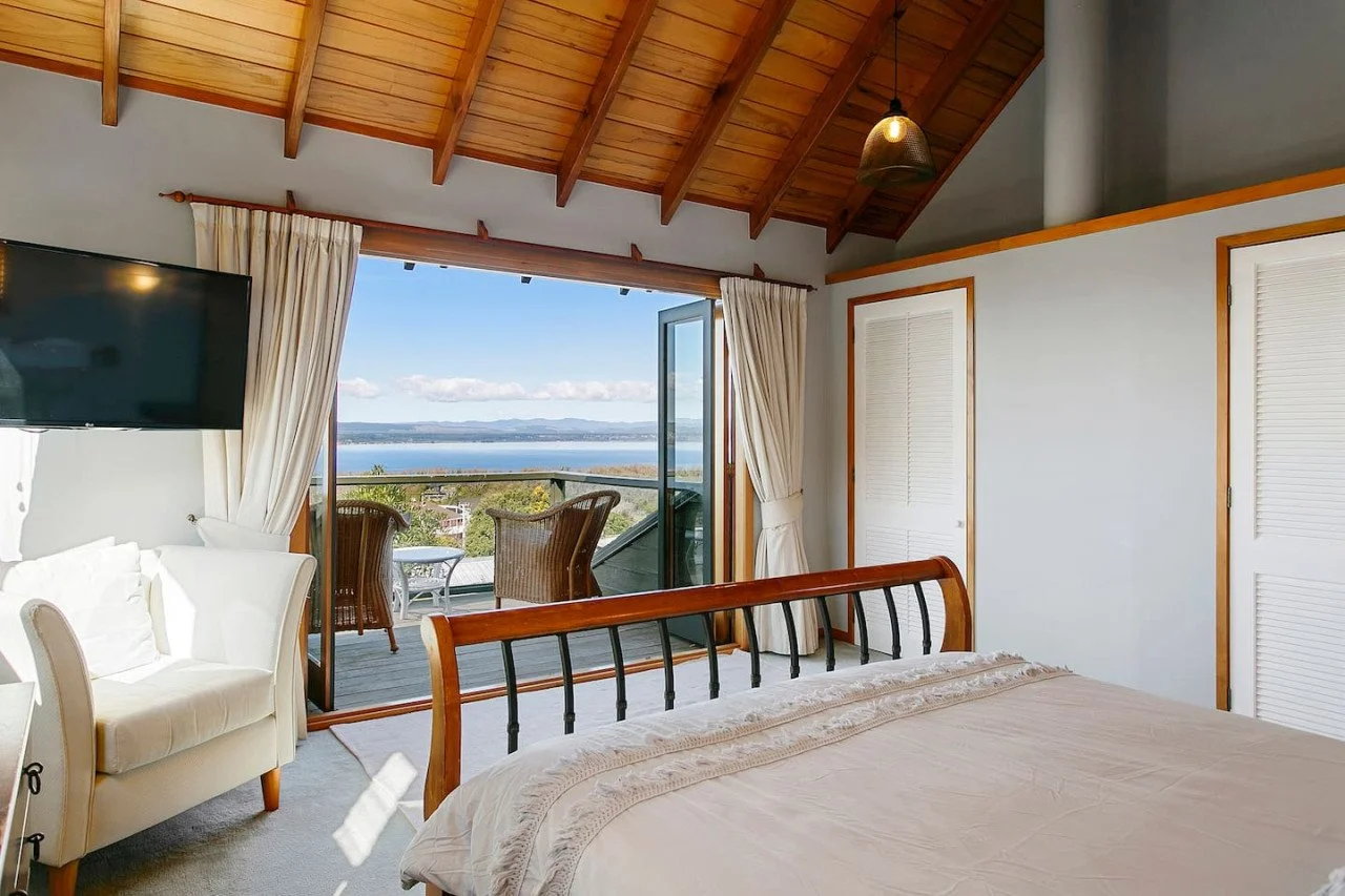 Bedroom with a sloped wooden ceiling, a large open door leading to a balcony with outdoor seating, overlooking water and distant hills, with cream-colored walls, a bed with a wooden and metal headboard, a white armchair, a television mounted on the w