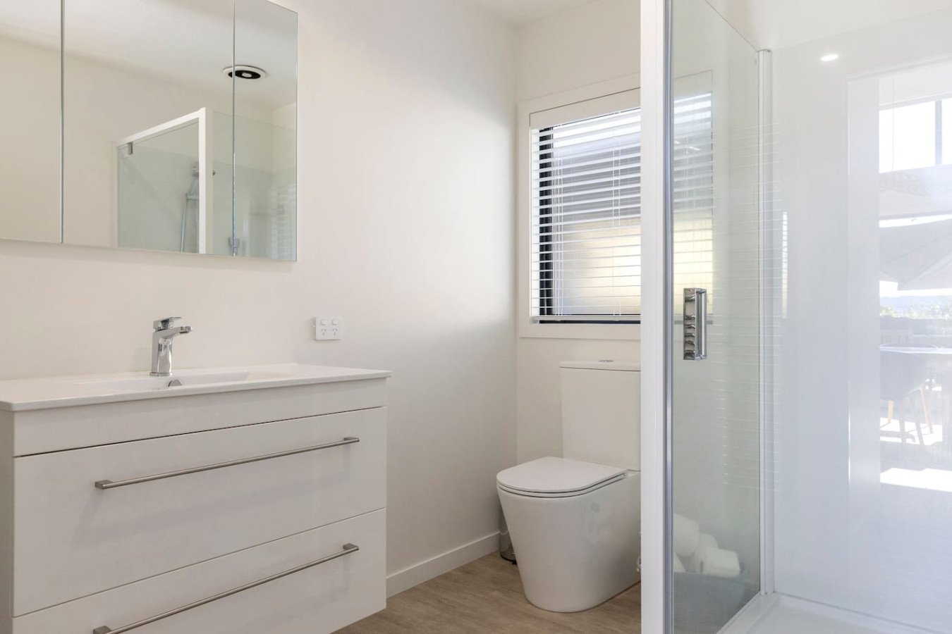A modern, minimalist bathroom with a white vanity, a mirror, a wall-mounted toilet, window with blinds, and a glass shower enclosure.