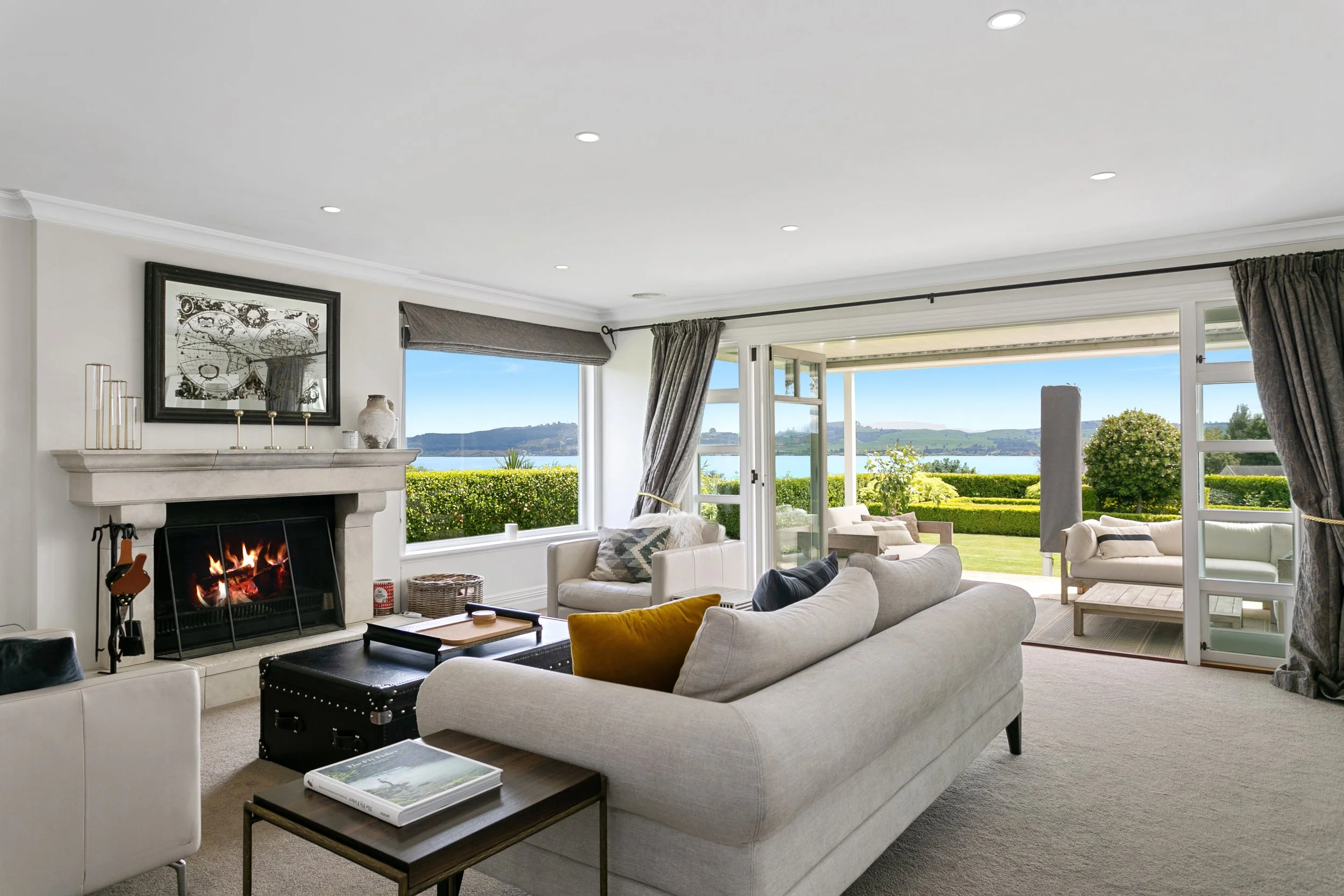 Living room with large windows and a view of a lake and greenery, fireplace, white sofas, and outdoor seating area.