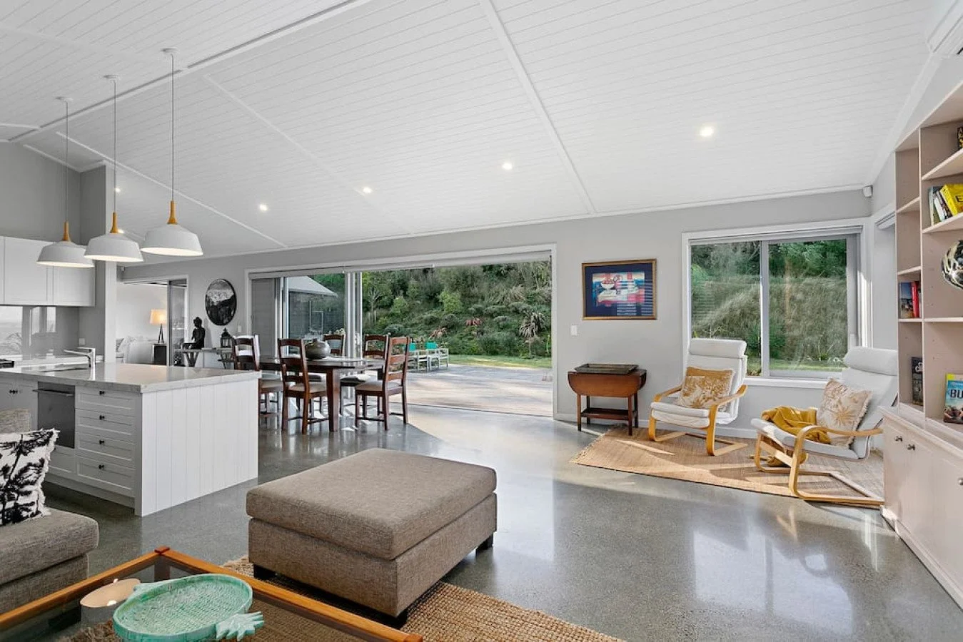 Spacious living room with large windows, white walls, hardwood chairs, a bookshelf, and a view of a backyard garden.