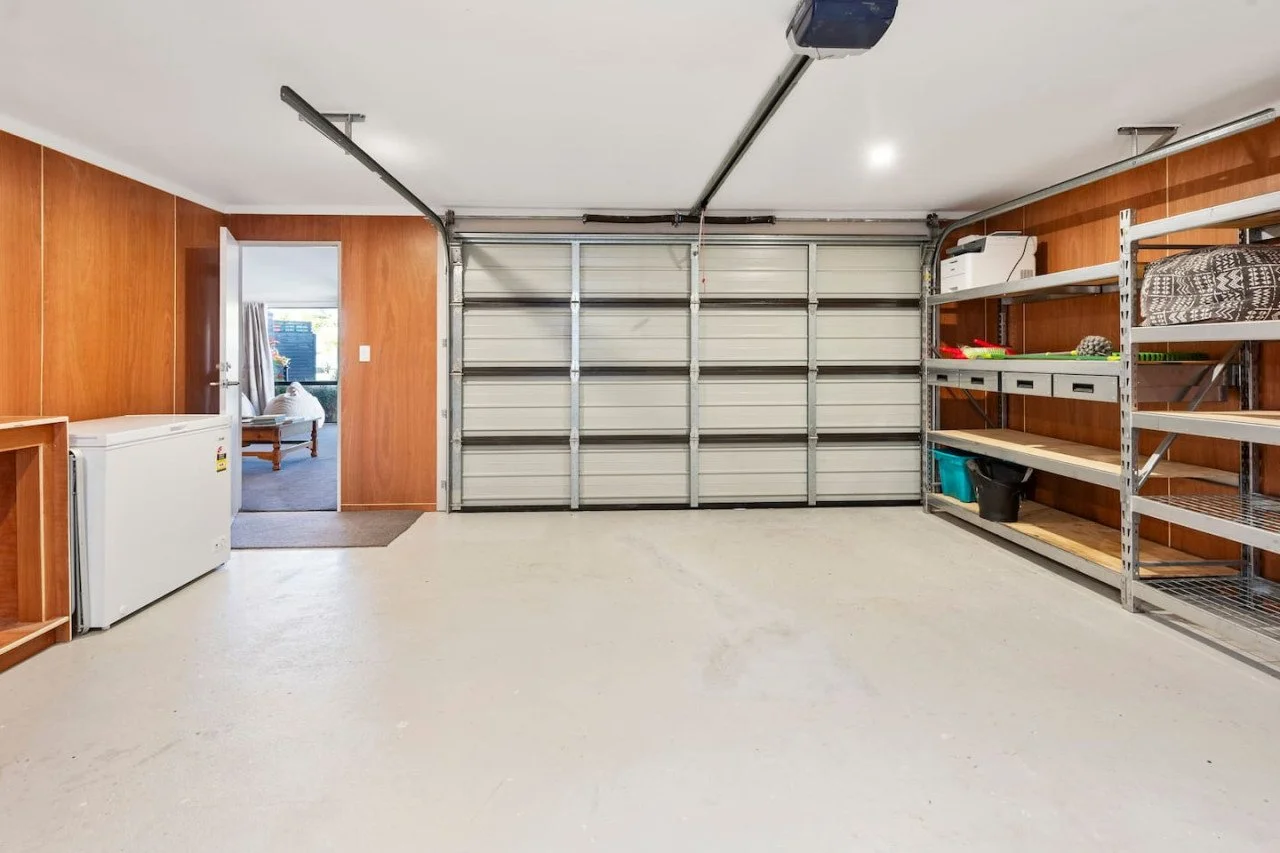 Empty garage with white overhead door, wooden wall paneling, metal shelving, and a small chest freezer.
