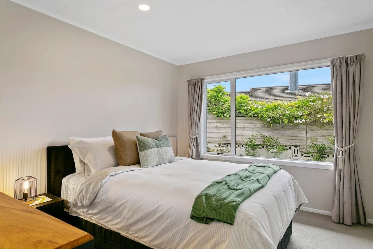 Clean bedroom with white bed, green blanket, pillows, bedside lamp, window with curtains, outside greenery, and a wooden dresser.