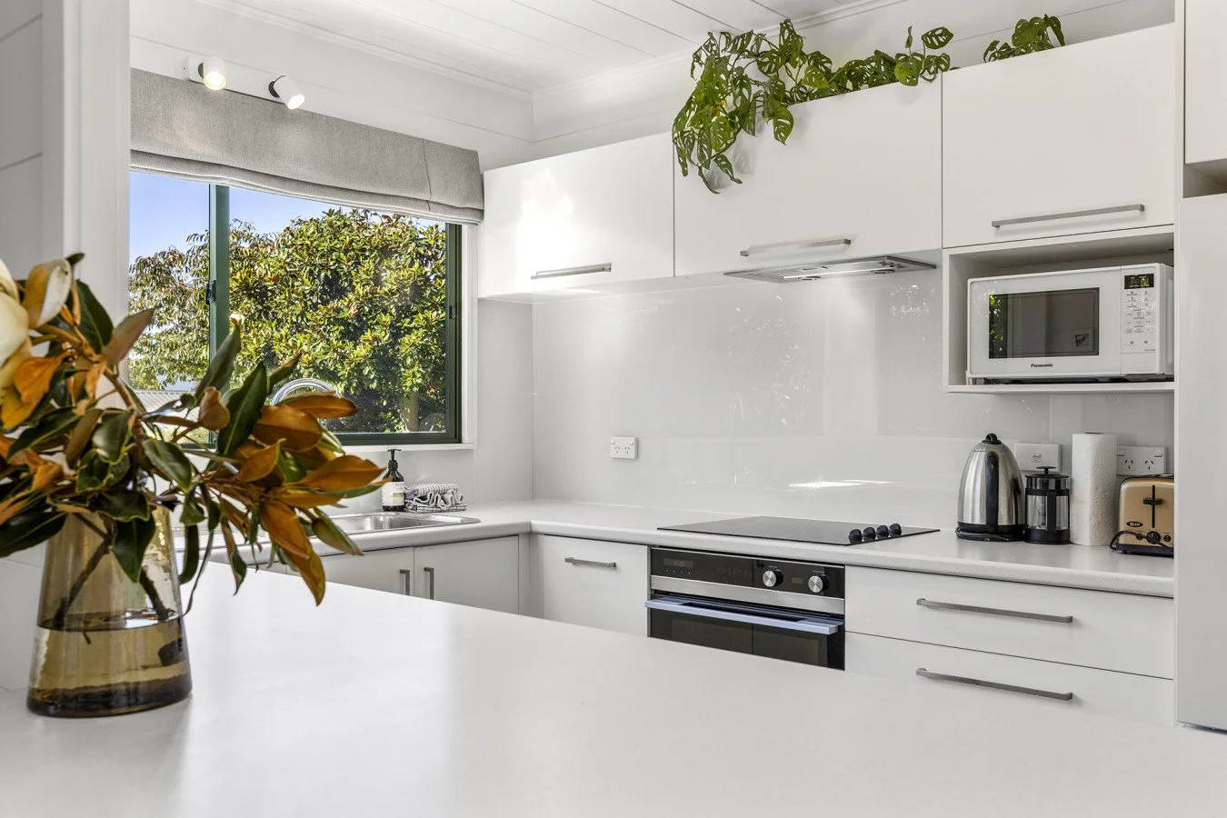 Modern white kitchen with a window showing trees outside, a vase with flowers on the counter, and appliances including a microwave, kettle, and toaster.