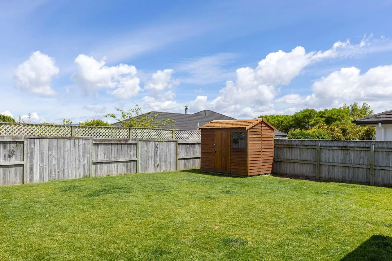 Backyard scene with a wooden shed, a grassy lawn, and a weathered wooden fence under a partly cloudy sky.