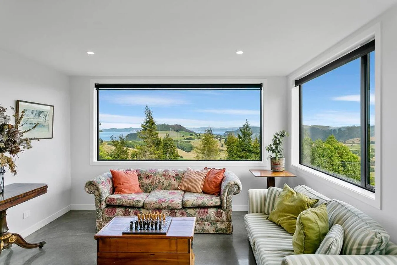 Living room with large windows showcasing a scenic view of trees, hills, and water, decorated with floral and striped sofas, pillows, a chessboard on a wooden coffee table, and potted plants.