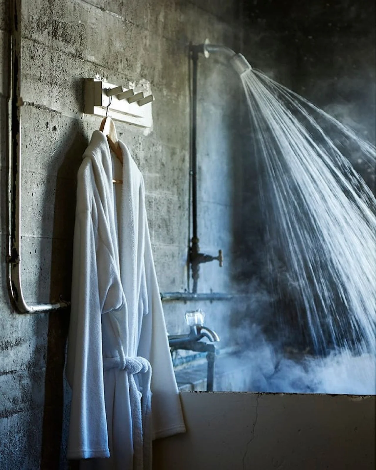 A shower area with an open showerhead producing a stream of water, a concrete wall, and two white bathrobes hanging on a wooden hook.