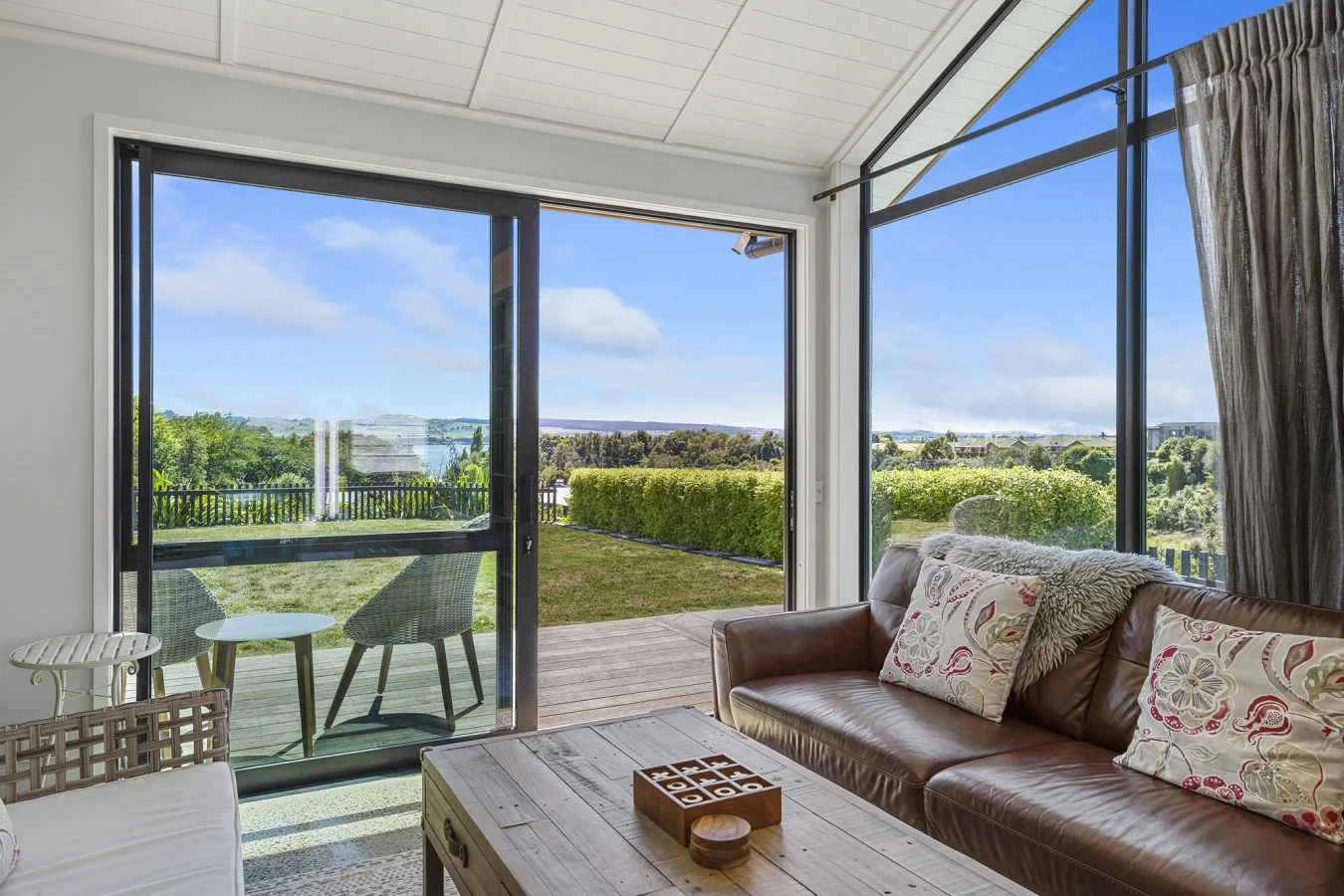 Living room with large glass sliding doors opening to a deck and a view of green hills and trees under a blue sky.