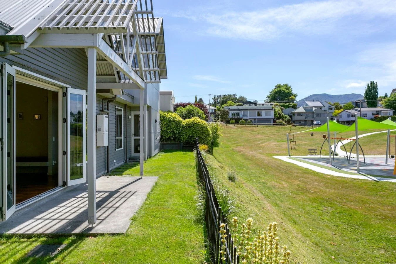 View of a house exterior with a small patio, surrounded by a green lawn and a black metal fence, with a park area featuring shade structures, a picnic table, and a playground, under a clear blue sky.