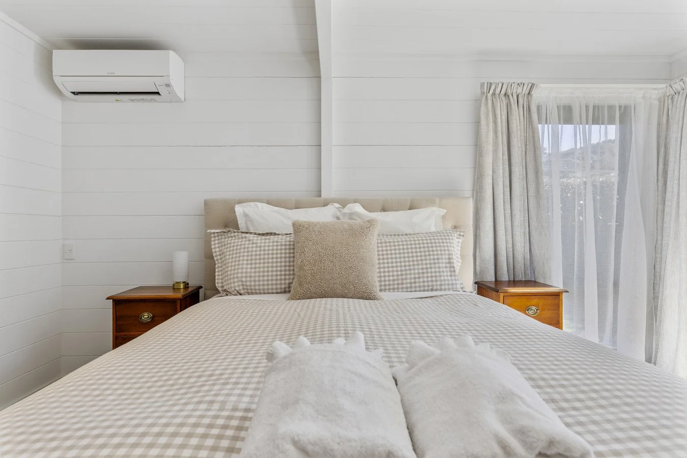 A neatly made bed with checkered bedding, beige pillows, and white towels in a bright bedroom with white paneled walls, two wooden nightstands, and sheer curtains covering a window.