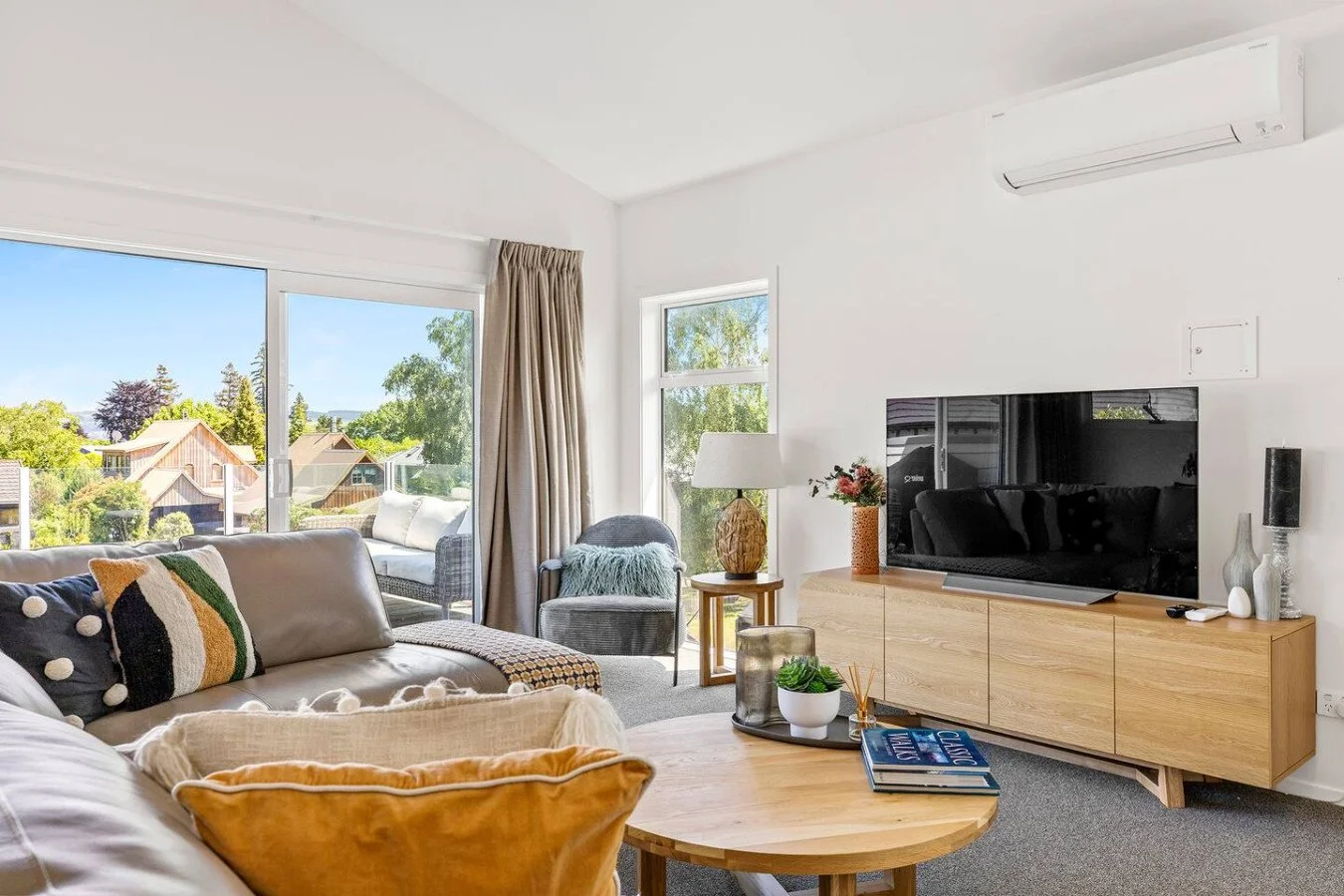 Bright living room with large windows, a beige sofa with colorful pillows, a coffee table with magazines, a TV on a wooden stand, and a side table with a lamp and a flower vase.