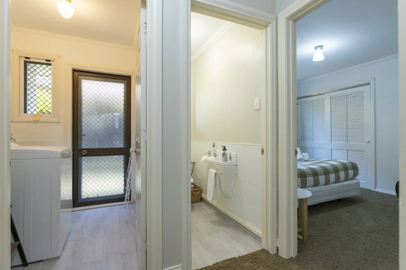 View of three connected rooms, including a laundry area with a washing machine and window, a bathroom with a wall-mounted sink, and a bedroom with a bed and closet.