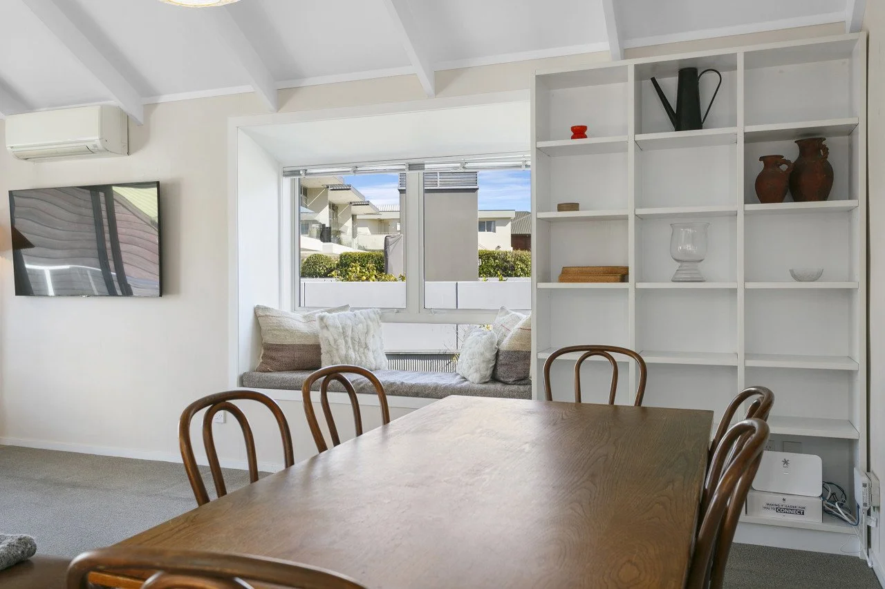 Dining room with wooden table and four chairs, white wall with shelves holding decorative items, window seat with pillows, wall-mounted television, air conditioning unit.