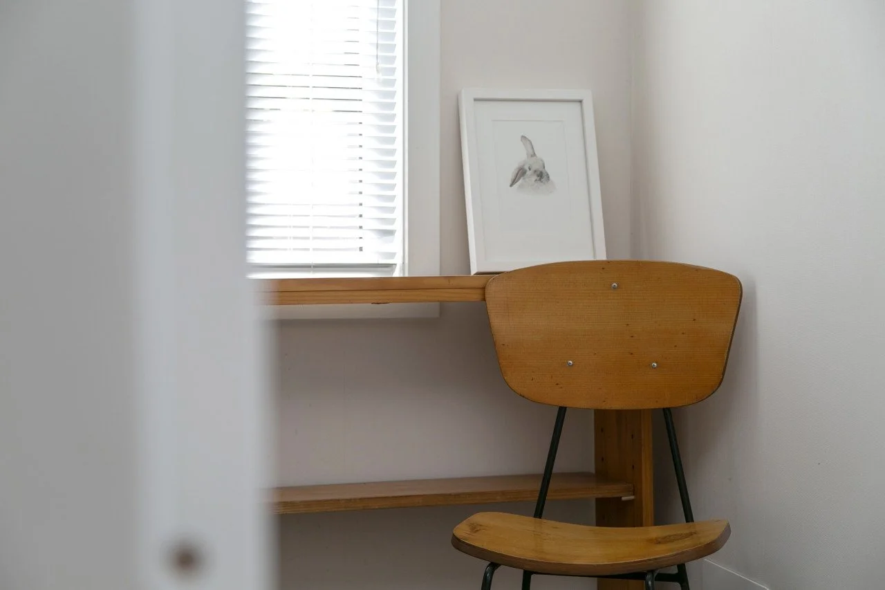 A cozy corner with a wooden desk, a framed art print of a rabbit, and a wooden chair in front of a window with white blinds.