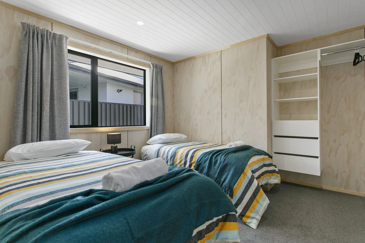 A bedroom with two beds, each with striped bedding and a rolled towel, a window with gray curtains, a black nightstand with a lamp between the beds, and an open closet with shelves. The walls are wood paneling, and the ceiling is white.