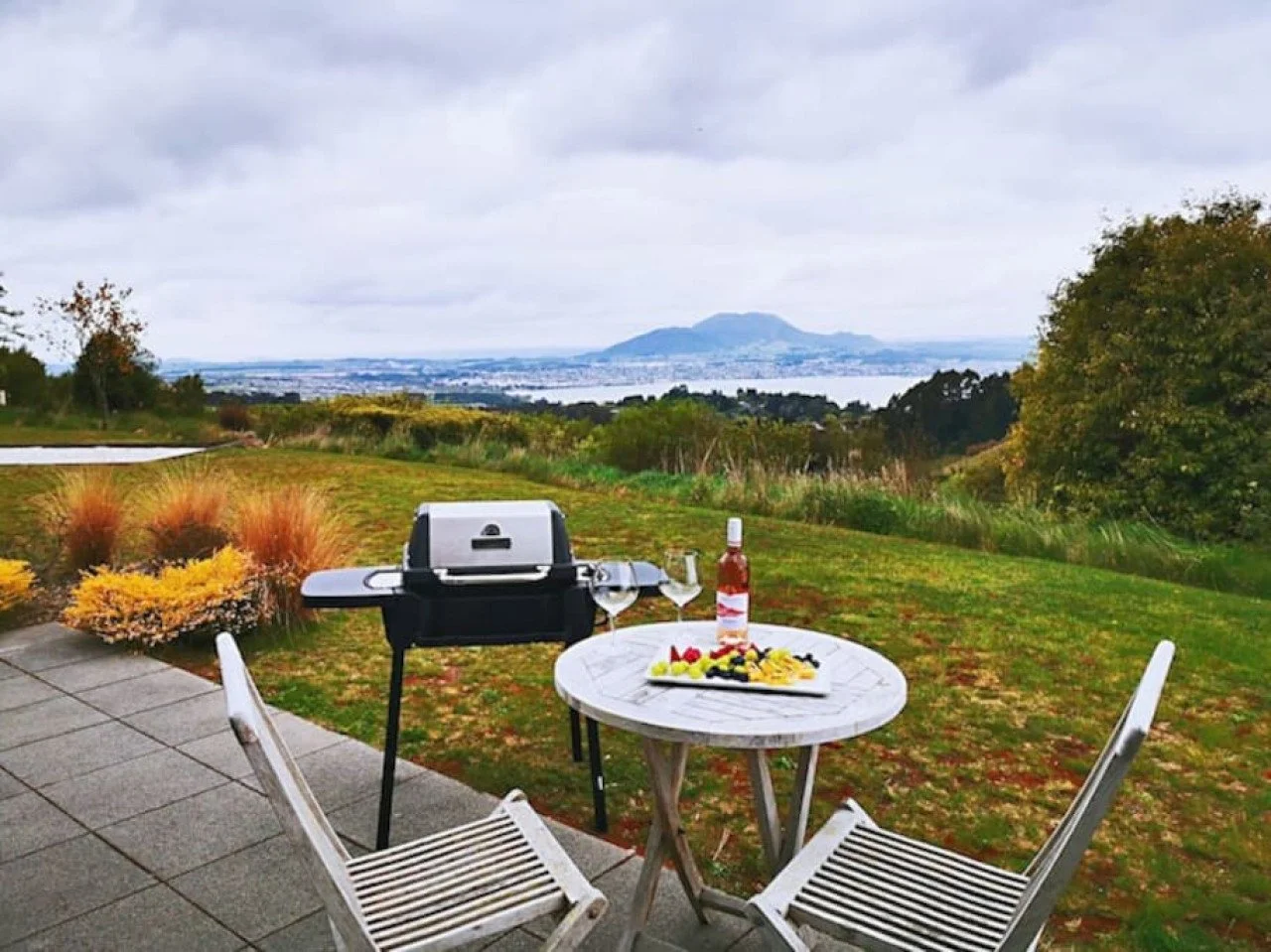 Outdoor patio with a small round table and two chairs, a grill, a bottle of hot sauce, a wine glass, and a platter of food, overlooking a green landscape with trees, shrubs, and a distant mountain under cloudy sky.