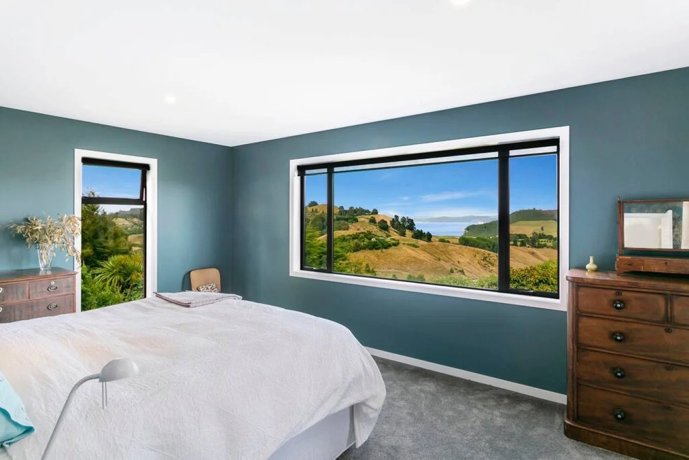 Bedroom with blue walls, large window showing a scenic landscape of rolling hills and ocean, wooden dresser, and bed with white linens.