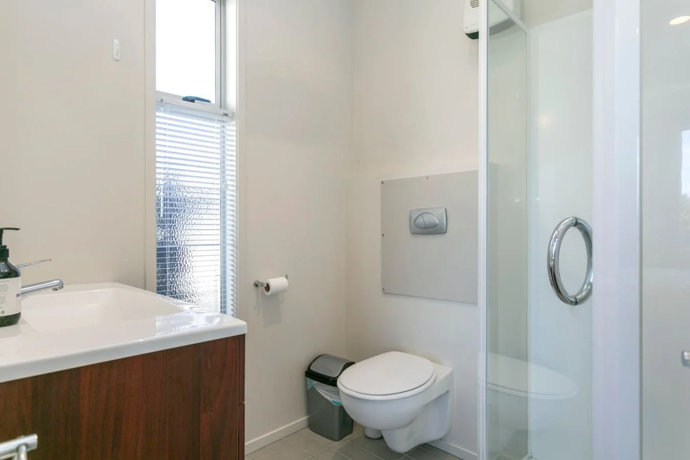 Bathroom with a toilet, a small window, a sink with soap, a trash bin, a paper towel holder, and a glass shower enclosure.