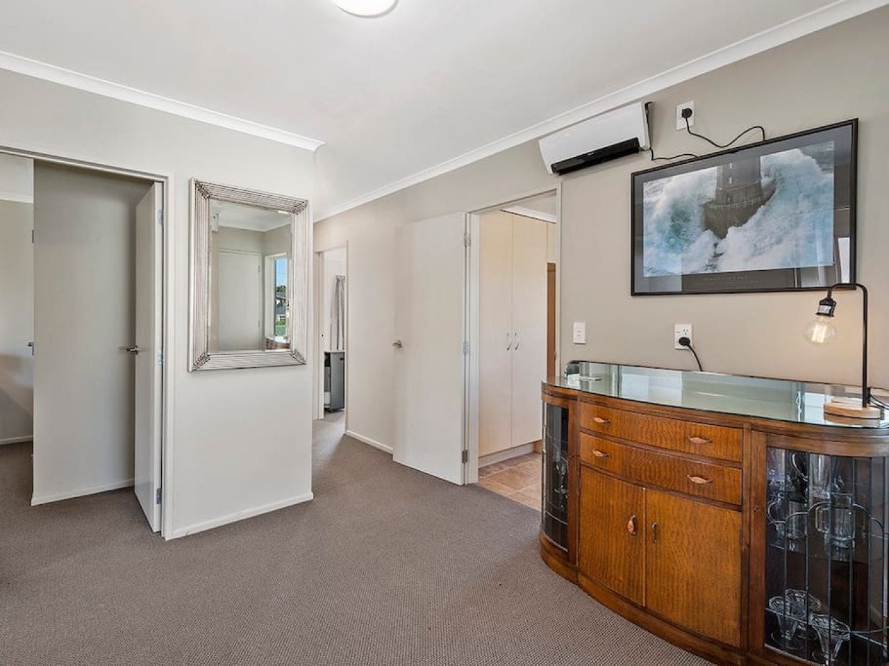 Living room with beige walls, a wall-mounted TV showing a lighthouse, a wooden cabinet with glass doors, a corner desk lamp, an air conditioning unit, and open doors leading to other rooms.