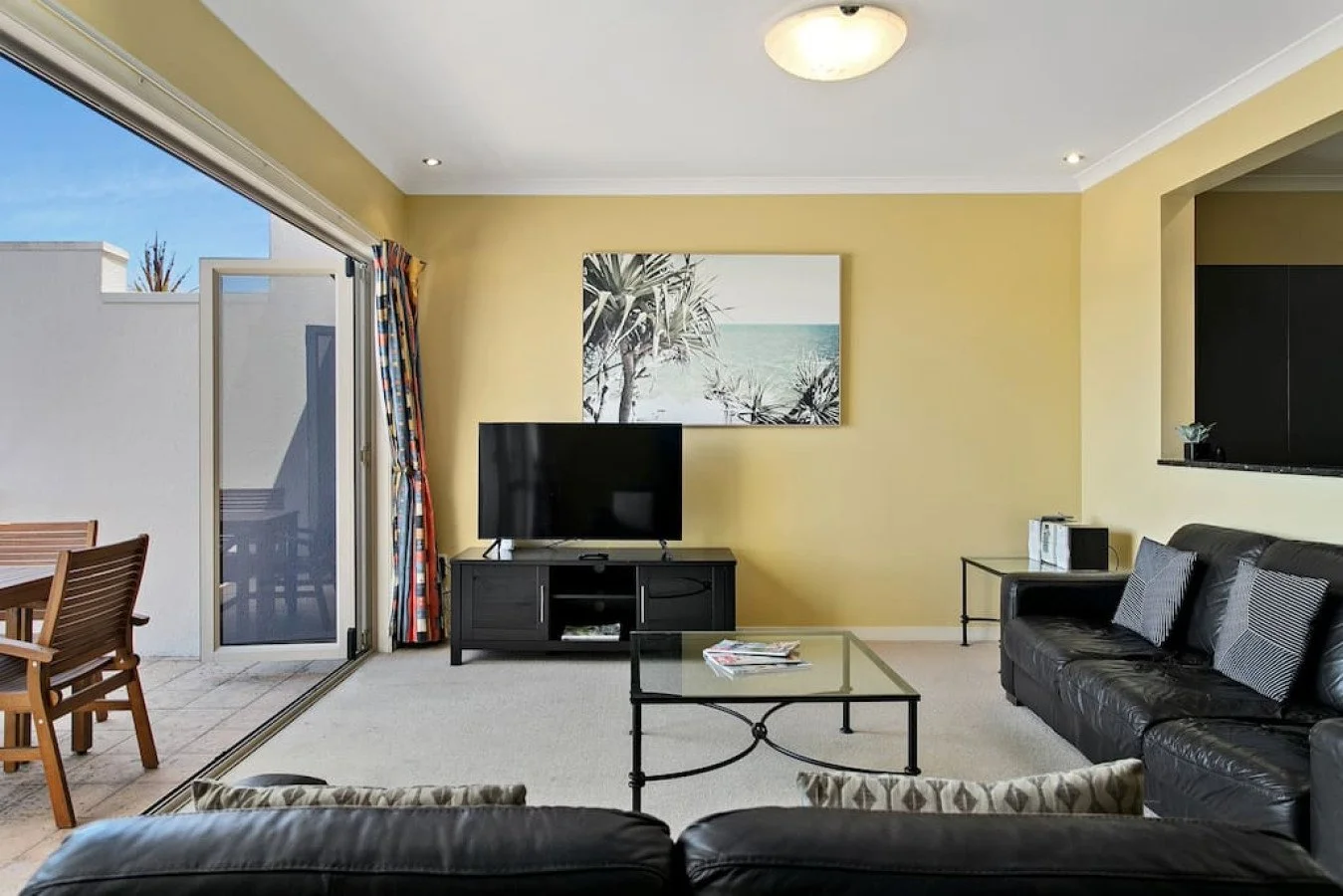 Living room with yellow walls, black sofa, glass coffee table, TV on black stand, wall art of trees and beach, sliding glass door with outdoor patio and wooden table and chairs, curtains, carpeted floor.