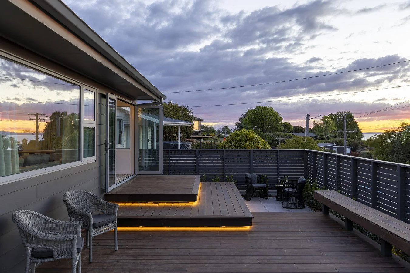 View of a wooden deck with outdoor furniture, including two wicker chairs and three black chairs with a small table, overlooking a landscape with trees and a sunset sky.