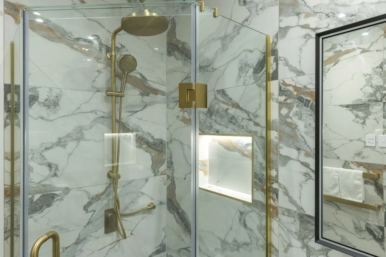 Modern bathroom with a marble wall shower enclosure featuring gold fixtures, a glass door, and a window, with a black-framed mirror and a towel rack.