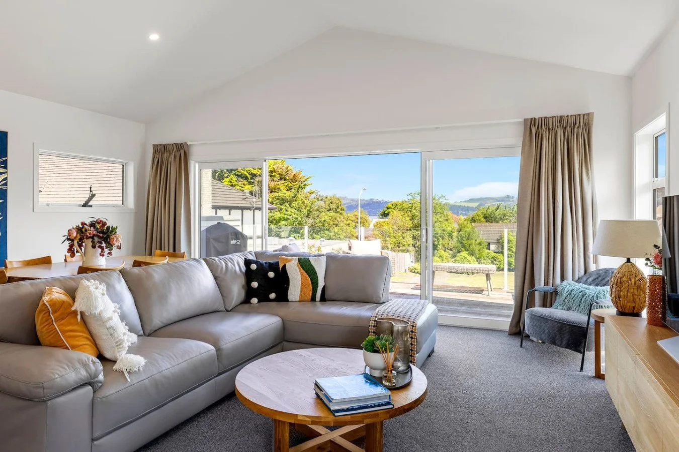 Living room with a large light gray sectional sofa, colorful throw pillows, a round wooden coffee table with books and decor, a sliding glass door leading to an outdoor deck with views of trees, houses, and distant hills, beige curtains, a patterned 