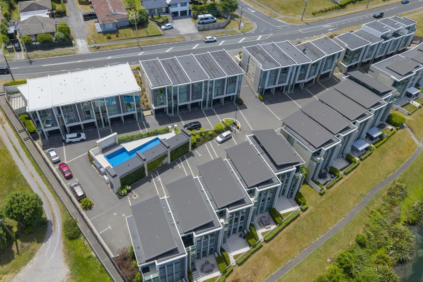 Aerial view of modern residential townhouses with gray roofs, small front yards, and parking lots. There is a swimming pool enclosed by fencing in the center of the complex, surrounded by lush greenery and a walking path.