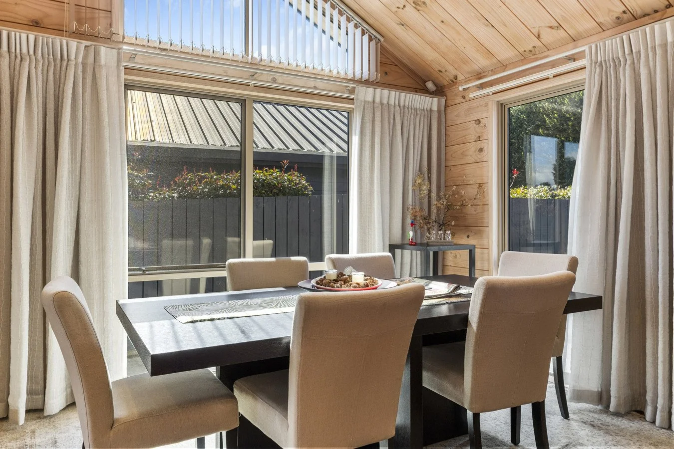 Dining room with a black rectangular table surrounded by six beige upholstered chairs, decorated with a centerpiece of candles and pinecones, with large windows, cream curtains, wood-paneled walls, and greenery outside.
