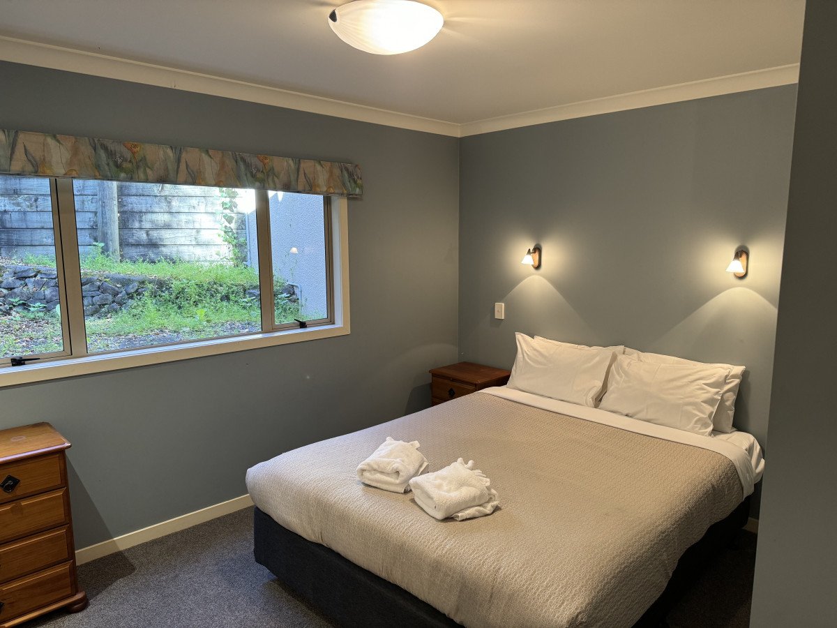 Hotel room with a bed, two pillows, folded towels, wooden nightstands, wall-mounted lights, window with a view of greenery, and a ceiling light.