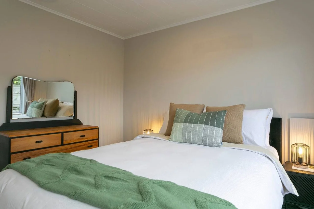  slaapkamer met wit beddengoed, groene en beige kussens, een groene deken, een houten dresser met mirror, nachtkastje met een lampje, en een raam met gordijnen.