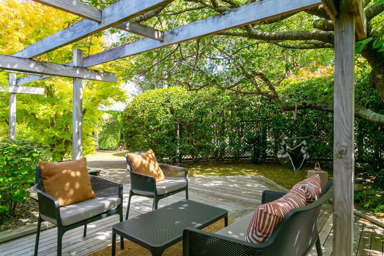 Outdoor patio area with black metal chairs, tan cushions, and striped pillows under a wooden pergola, surrounded by lush green trees and bushes, with a hanging swing seat in the background.