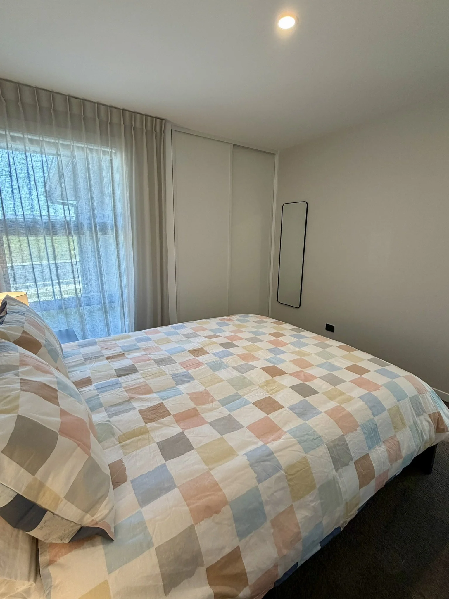 A bedroom with a large bed covered in a pastel-colored checkered quilt, two matching pillows, a window with sheer curtains, a cabinet with sliding doors, and a narrow mirror mounted on the wall.