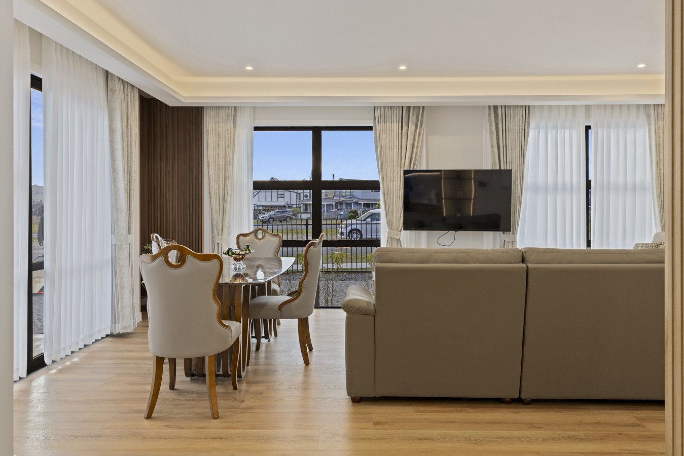 Living room with beige sofa, dining table with chairs, large windows with curtains, and a mounted flat-screen TV.