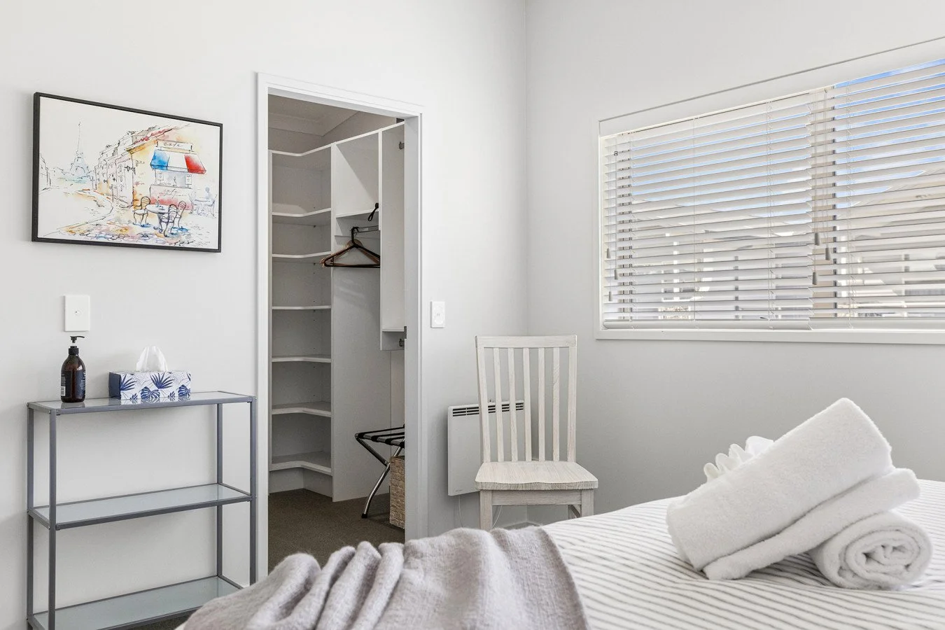 A bedroom with white walls, a bed with rolled white towels, a chair, a window with blinds, an art piece on the wall, and an open closet with shelves and hangers.