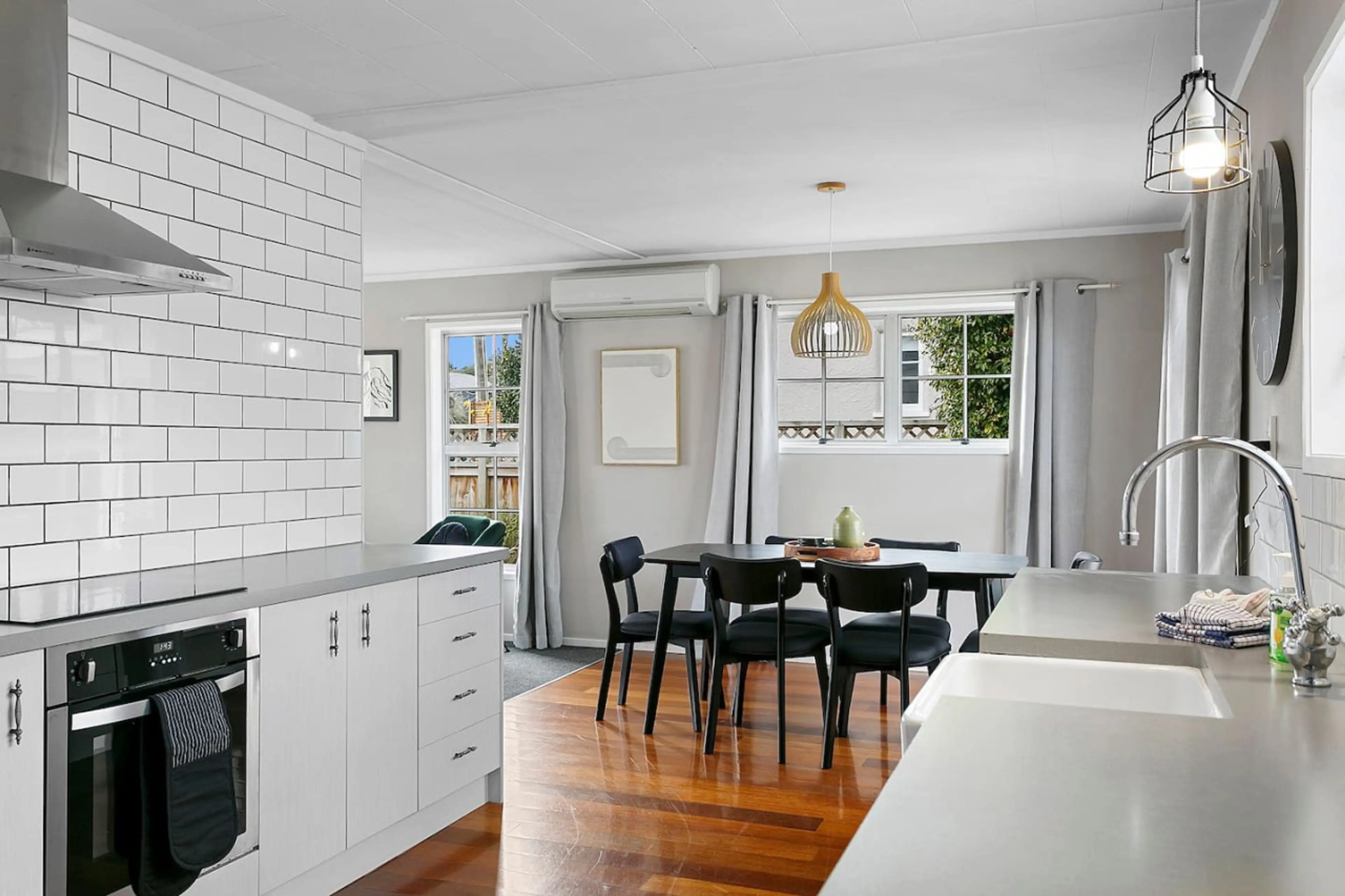 Bright kitchen and dining area with white cabinets, a black dining table with black chairs, white curtains, and large windows with views of greenery.