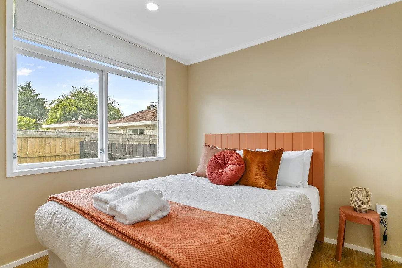 A bedroom with a bed that has a peach-colored headboard, white bedding, and decorative pillows in shades of orange and red. A folded towel is on the bed, and a small peach-colored side table with a lamp is beside it. A large window shows a view of a 