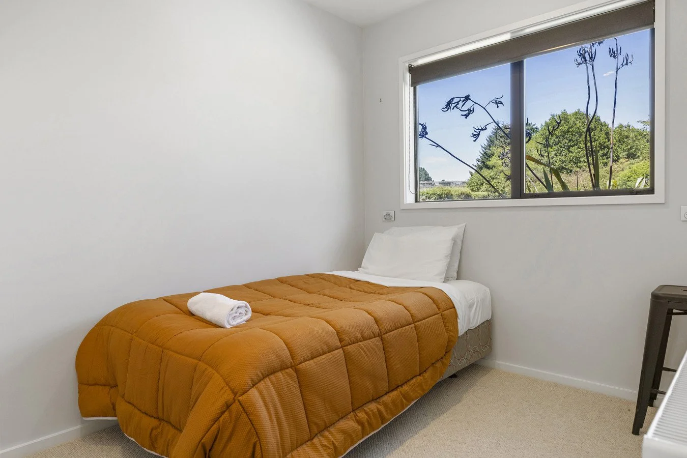 A simple bedroom with a single bed covered with an orange-brown quilt, a white pillow, and a rolled towel on top. There is a window with a view of trees and blue sky, and a small dark-colored table in the corner.
