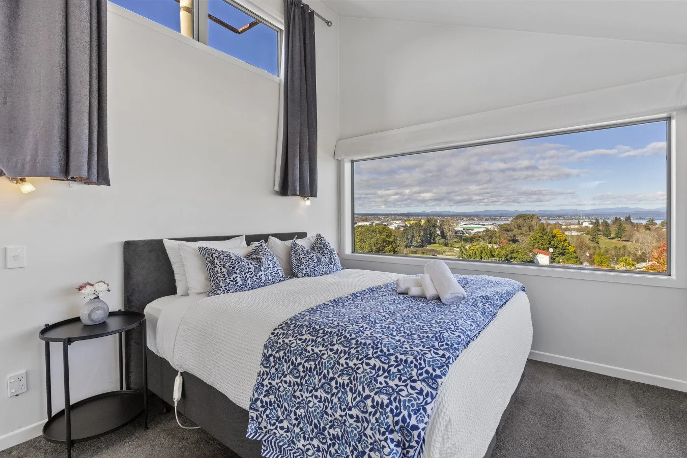 A bedroom with a large window showing a scenic view of trees, water, and sky. The bed has white bedding with blue patterned pillows and a matching blue throw. A small black nightstand with a vase and flowers is next to the bed.