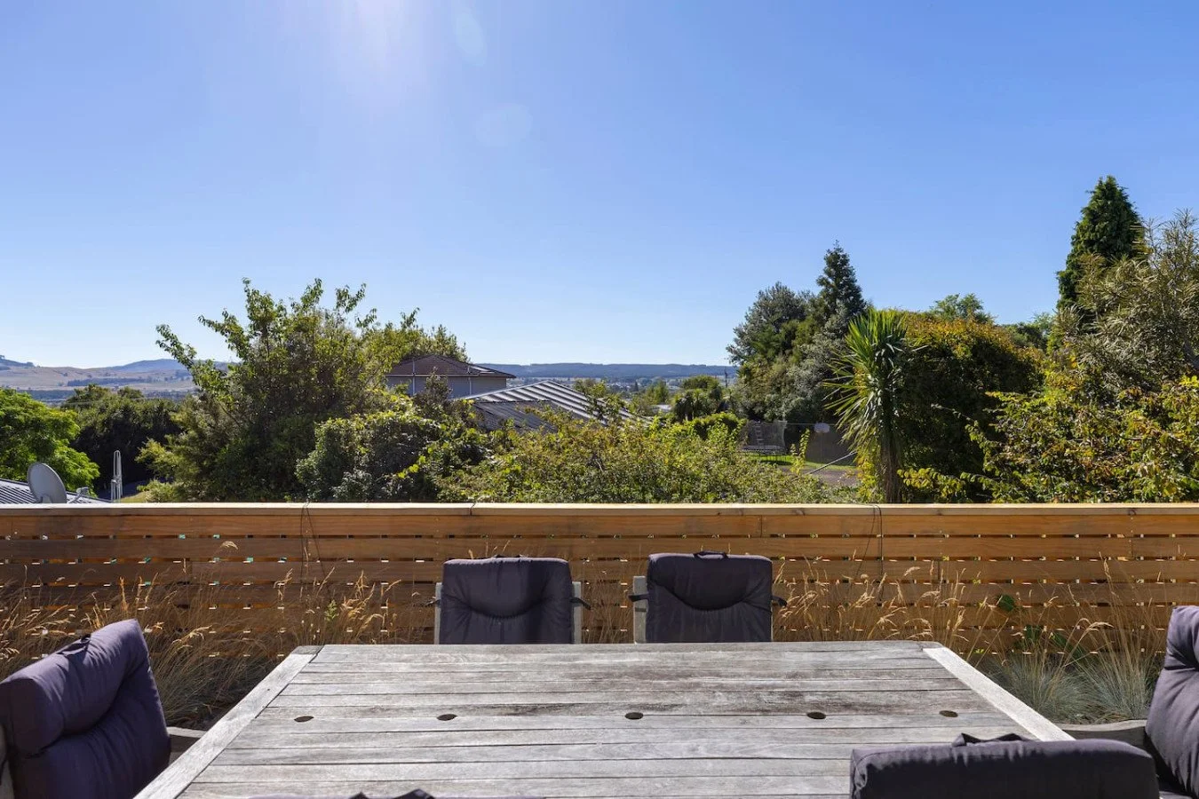 A patio with a wooden table and chairs, overlooking a lush backyard with trees and a clear blue sky.
