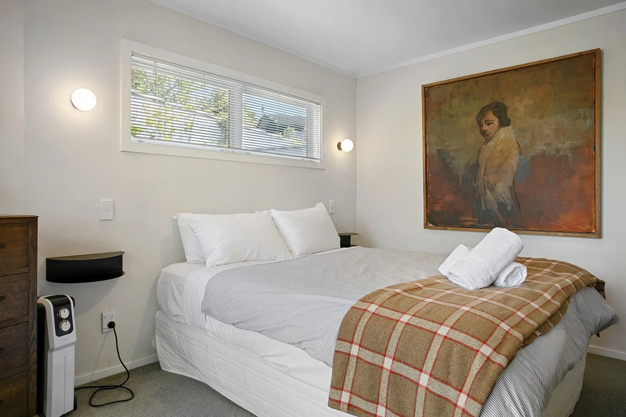 A neatly made bed with white pillows and bedding, a brown plaid blanket draped over the foot, towels on top, and a large framed painting of a woman on the wall behind. There are two wall-mounted lamps, a window with blinds, and a small black side tab